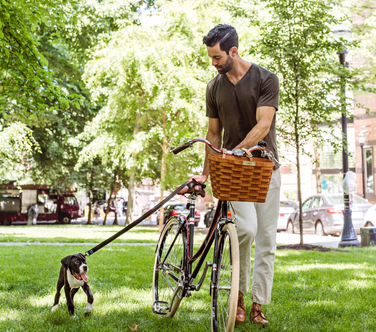 A man walking his bike through a park with his puppy.