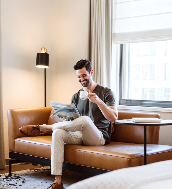 A man on a couch reading a paper and drinking a coffee in his suite at the Heathman Hotel in Portland, Oregon.