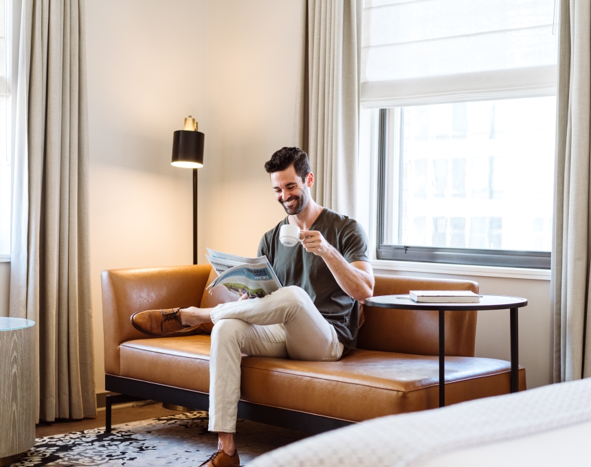 A man on a couch reading a paper and drinking a coffee in his suite at the Heathman Hotel in Portland, Oregon.