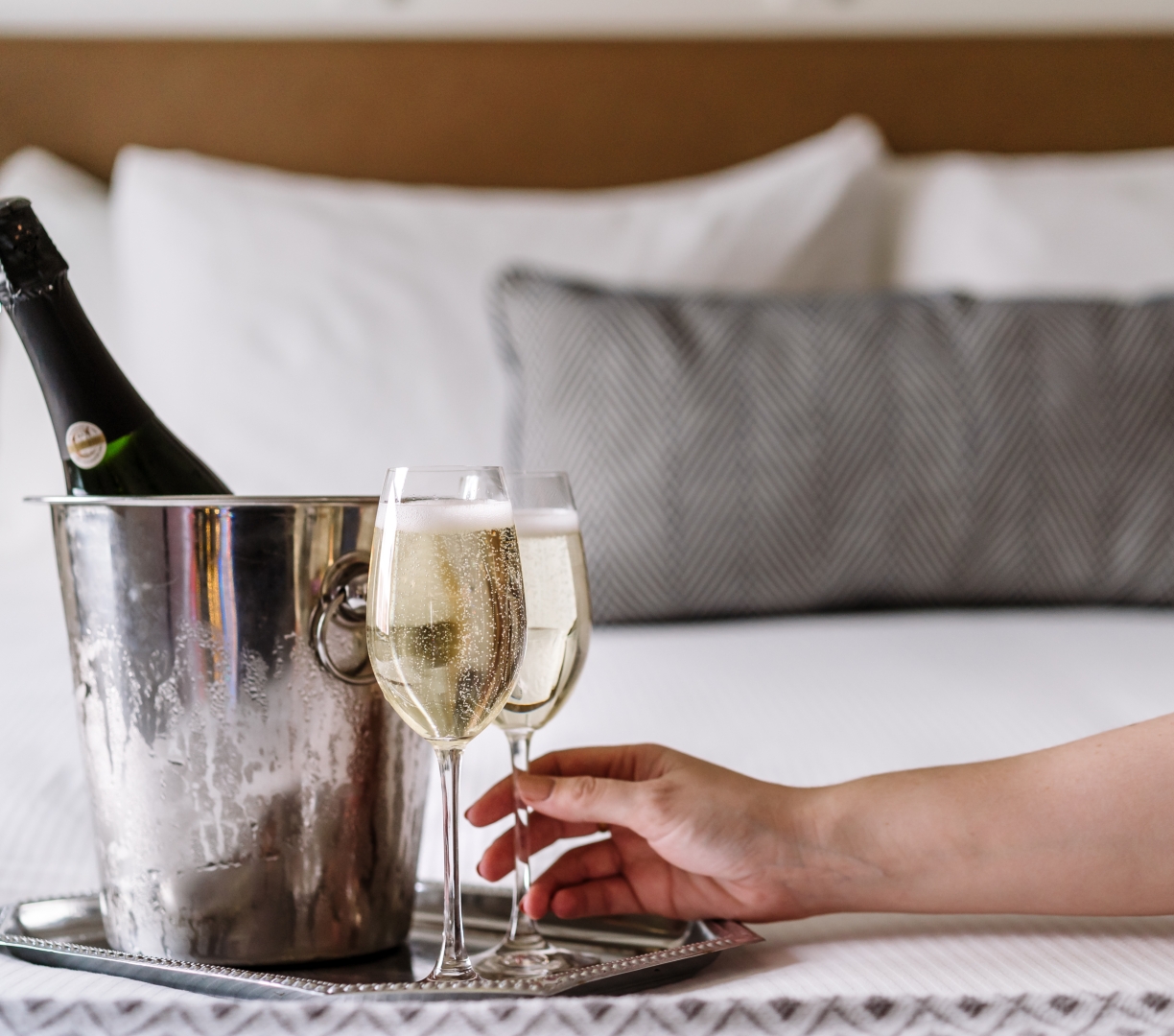 A silver tray with glasses and a bottle of champagne in an ice bucket on a hotel bed.