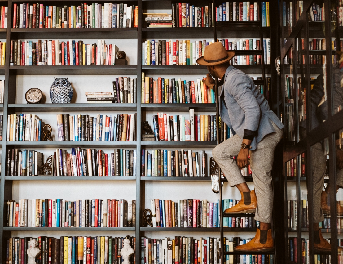 A man on a ladder choosing a book from a tall library shelf.