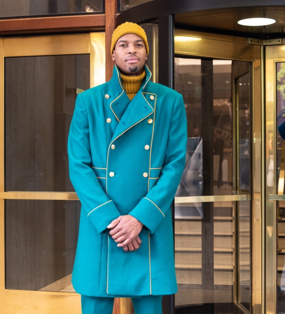 A man in a long blue coat and a yellow beanie standing in front of the Heathman Hotel in Portland, Oregon.