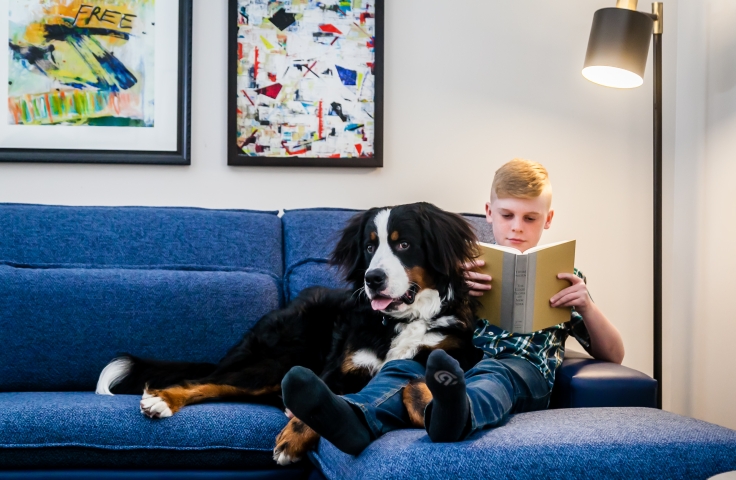 A young boy sitting with his dog on a couch and reading a book.
