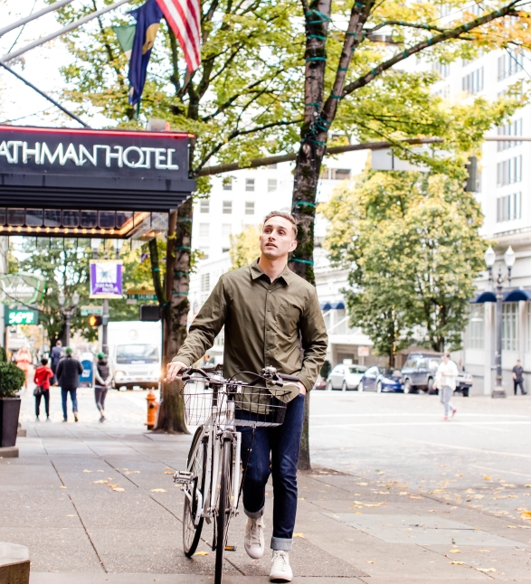 A man walking his bike on the sidewalk by the Heathman Hotel.