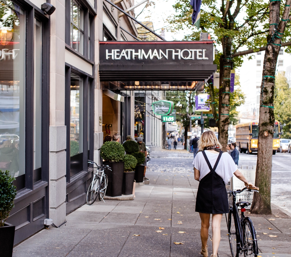 A woman walking a bike on the sidewalk by the Heathman Hotel.